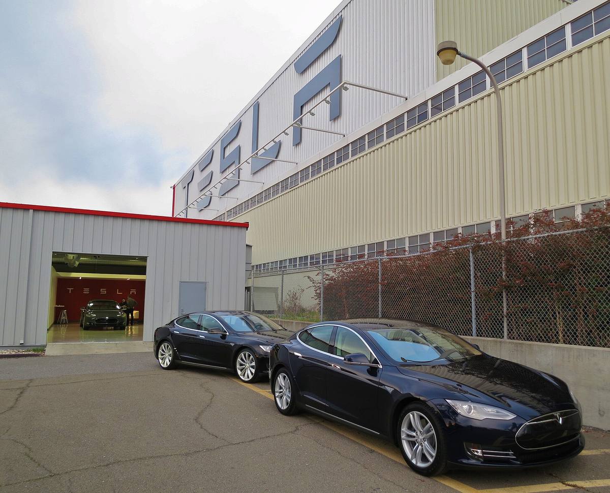 Dvě tmavě modré Tesly Model S zaparkované před halou s nápisem Tesla