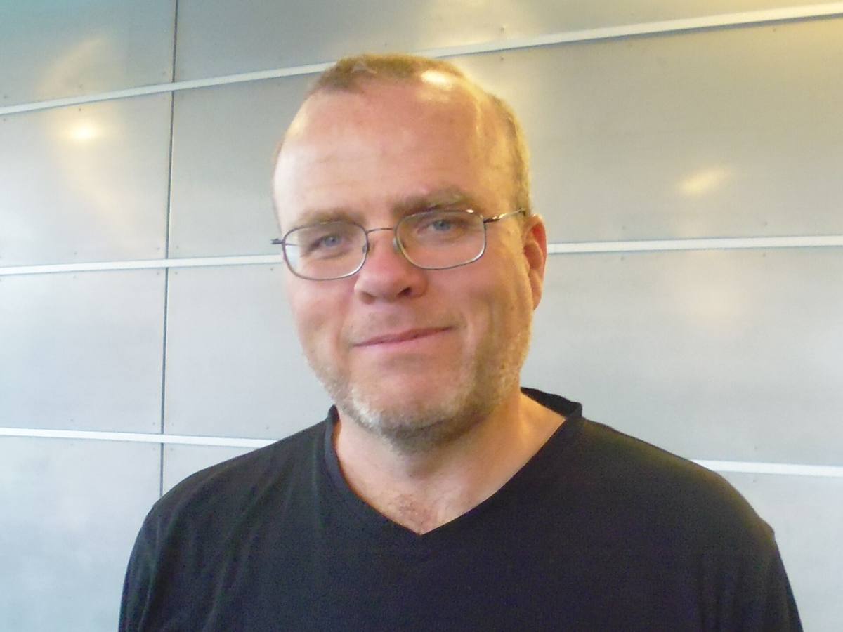 Rasmus Lerdorf s brýlemi a v černém tričku před světlou stěnou