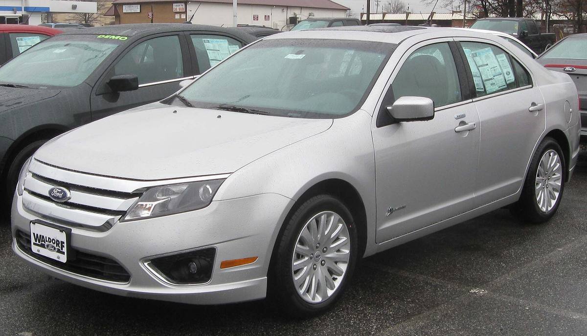 Stříbrný sedan Ford Fusion Hybrid ročníku 2010 na mokrém parkovišti prodejce