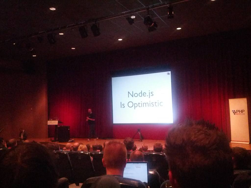 Promítací plátno s nápisem Node.js Is Optimistic v sále plném posluchačů
